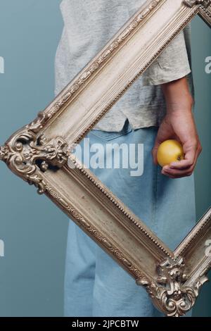 Ein junger Mann aus dem Jahrtausend hält einen Apfel auf der Handfläche in einem vergoldeten Bilderrahmen Stockfoto