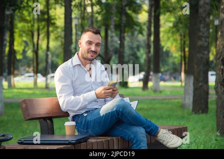 Ein lächelnder Mann sitzt auf einer Bank im Park und zählt den Gewinn. Ein junger Mann auf einem Hintergrund von grünen Bäumen, ein heißer sonniger Sommertag. Warmes, weiches Licht, Nahaufnahme. Stockfoto