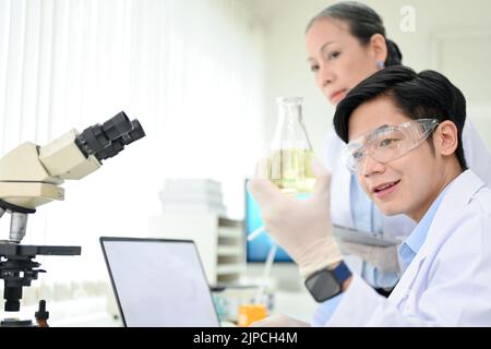Ein hübscher, intelligenter junger asiatischer Wissenschaftler oder Chemiker, der mit seiner leitenden weiblichen Vorgesetzten im Wissenschaftslabor ein chemisches Experiment unternimmt. Stockfoto