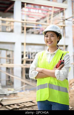 Porträt einer attraktiven und selbstbewussten jungen asiatischen Ingenieurin in Uniform und weißem Hardhut, die auf der Baustelle steht. Stockfoto