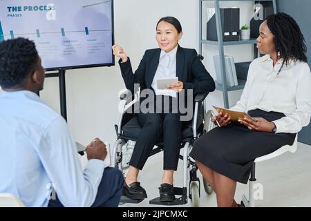 Asiatische Geschäftsfrau mit Behinderung, die ihren Kollegen Geschäftsberichte erstattet, während sie ihr während der Präsentation im Büro zuhört Stockfoto