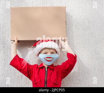 Neukäufe oder Lieferung aus dem Geschäft. Kleinkind Junge in roten Jumpsuit, Weihnachtsmütze. Auf dem Gesicht ist blaue medizinische Maske mit Lächeln auf sie gemalt. Hält Pappe Stockfoto