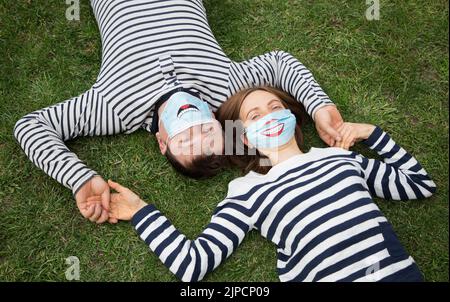 Verliebte Männer und Frauen in gestreiften Pullovern liegen auf dem Gras und halten sich die Hände. Beide tragen blaue medizinische Masken mit einem lustigen Lächeln auf sie gemalt. Positiv, Stockfoto