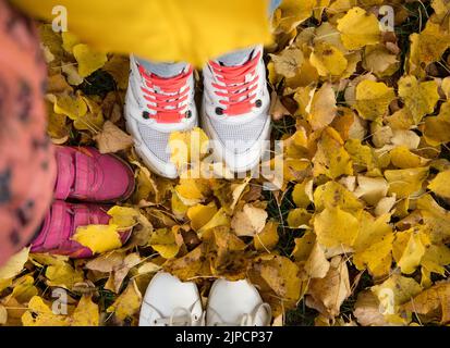 Herbstliche Trockenblätter auf dem Boden mit den Füßen der Menschen in Stiefeln und Turnschuhen. Draufsicht von oben, Herbstkonzept-Kulissen. Saisonale Atmosphäre Stockfoto