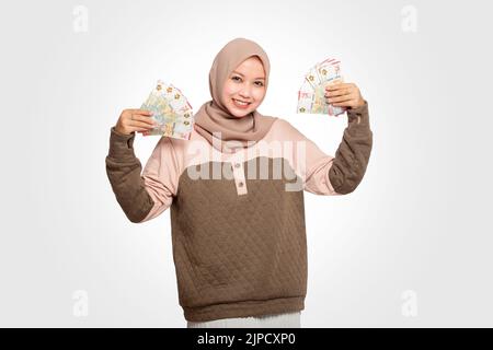 Porträt einer glücklichen asiatischen muslimischen Frau, die Rupiah-Geld hält. Währung von 75000 Rupiah am 75.. Tag der Unabhängigkeit Indonesiens. Stockfoto