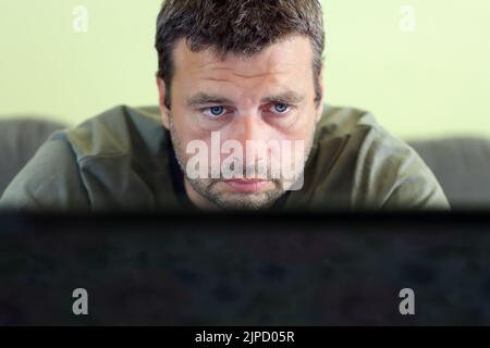 Nahaufnahme eines Kopfporträts eines weißen kaukasischen bärtigen Mannes mit hübschen blauen Augen, der von zu Hause aus auf einem Laptop arbeitet. Selektiver Fokus Stockfoto