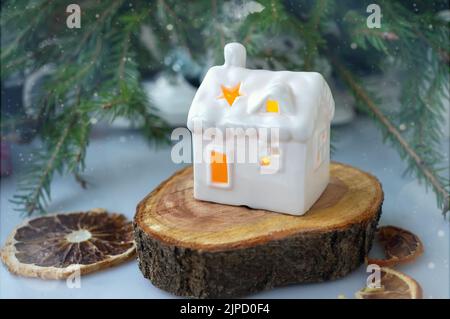 Weihnachtsdekor. Kiefernzweige, eine Girlande und eine getrocknete Orange und eine keramische Lampe in Form eines weißen Keramikhauses Stockfoto