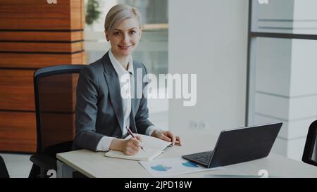 Porträt einer attraktiven blonden Geschäftsfrau, die am Tisch sitzt und in einem Notebook in die Kamera lächelt, im modernen Büro im Innenbereich Stockfoto