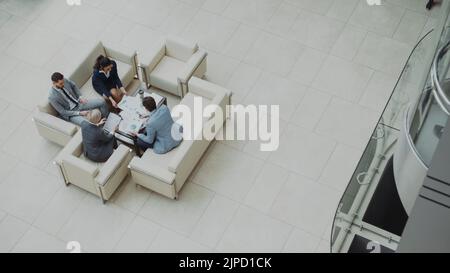 Draufsicht auf Kollegen aus der Gruppe von Geschäftsleuten, die Finanzkarten diskutieren, die auf Sofas in der Lobby im modernen Business Center im Innenbereich sitzen Stockfoto