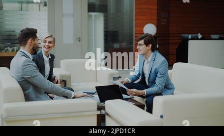 Fröhlicher Geschäftsmann, der Finanzberichte mit Geschäftspartnern diskutiert und auf dem Sofa in der modernen Bürohalle sitzt Stockfoto