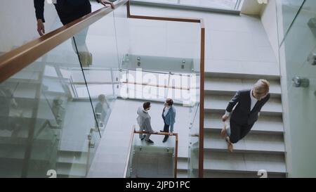 Blick von oben auf zwei Geschäftsleute treffen sich im modernen Bürozentrum im Treppenhaus und sprechen, während weibliche Kollegen Treppen steigen Stockfoto
