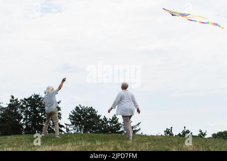 Ältere Frau in legerer Kleidung, die in der Nähe ihres Mannes auf einem grünen Hügel mit Drachen spielt Stockfoto