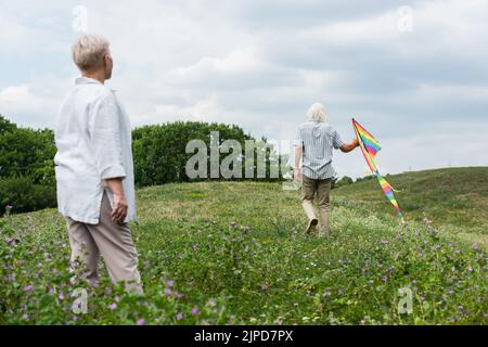 Rückansicht einer Frau mit grauem Haar, die den Ehemann ansieht, der mit dem Drachen auf einem grünen Hügel läuft Stockfoto