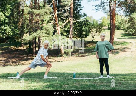 Der lange ältere Mann mit grauem Haar lächelt und macht Ausfallschritte auf der Fitnessmatte in der Nähe der Frau im Park Stockfoto