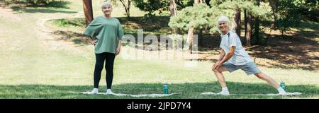 Ein langer älterer Mann mit grauem Haar lächelt und macht Ausfallschritte auf der Fitnessmatte in der Nähe der Frau im Park, Banner Stockfoto