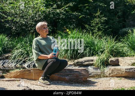 Rentnerin in Sportkleidung sitzt mit Sportflasche auf Holzbalken Stockfoto
