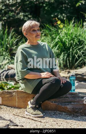 Rentnerin in Sportkleidung sitzt mit Sportflasche auf Holzbalken um grüne Pflanzen Stockfoto