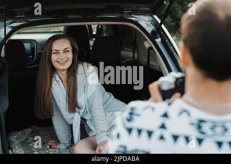 Fröhliche kaukasische Frau, die im Kofferraum eines SUV-Autos sitzt, während ihr Freund sie auf einer Kamera fotografiert Stockfoto