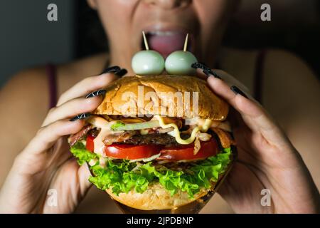 latina Mädchen beißen einen Hamburger sie hält mit beiden Händen. hamburger mit zwei Wachteleiern auf der Oberseite imitieren Augen. Street Food auf schwarzem Backgr Stockfoto