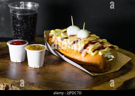 Hot Dog mit Saucen, Käse und zwei Wachteleiern auf einem Holztablett serviert und daneben zwei Plastikbecher mit Ketchup und Senf, neben Th Stockfoto