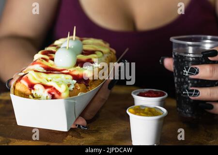 Mädchen hält in ihren Händen einen Hot Dog mit Saucen, Käse und zwei Wachteleiern auf einem Holztablett serviert und daneben sind zwei Plastikbecher mit Ket Stockfoto