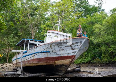 Drei kleine Jungen, die auf dem Bug eines verlassenen Fischerbootes sitzen. Sulawesi, Indonesien. Stockfoto