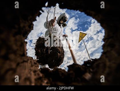 Blick von der Erdgrube auf die Person in Schutzanzug und Gasmaske, die in der Nähe des Warnzeichens für biologische Gefahrenstoffe steht, Schaufel in der einen Hand hält und in der anderen Bäume gegen den blauen Himmel mit weißen Wolken sättigen. Stockfoto