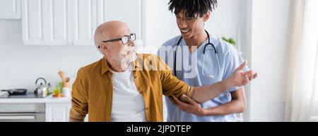 Positiver Rentner im Gespräch mit afroamerikanischer Krankenschwester zu Hause, Banner Stockfoto