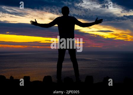 Eine nicht identifizierbare Silhouette eines Mannes steht mit ausgestreckten Armen vor einem bunten Abendhimmel. Stockfoto
