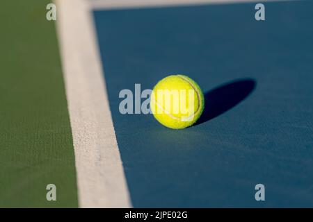 Gelber Tennisball in der Nähe der Grundlinie auf einem neuen blauen Tennisplatz mit Grün außerhalb der Grenzen. Stockfoto