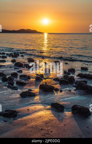 Eine untergehende Sonne über dem Horizont mit Hügeln im Hintergrund und Felsen und Kieselsteinen im Vordergrund an der Küste. Stockfoto