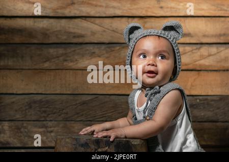 Schöne Baby Latina mit brauner Haut, mit Overalls und einer Mütze mit Mausohren, gestrickt mit der Häkeltechnik. Mit orangefarbenem Holzhintergrund A Stockfoto