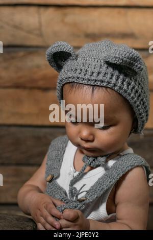 Wunderschönes, braunhäutig gebautes Latina-Baby, das mit den Händen abgelenkt ihren Overall hält und nach unten blickt. Mit einem orangefarbenen Holzhintergrund in einem armen Haus. Stockfoto