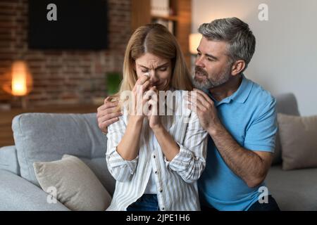Trauriger kaukasischer Mann mittleren Alters mit Bartumarmung, ruhig schreiende, unglückliche Frau im Wohnzimmer Stockfoto