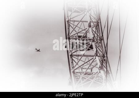 Ein monochromes Bild eines Flugzeugs, das am Himmel fliegt, mit einem Teil des London Eye im Vordergrund. Stockfoto