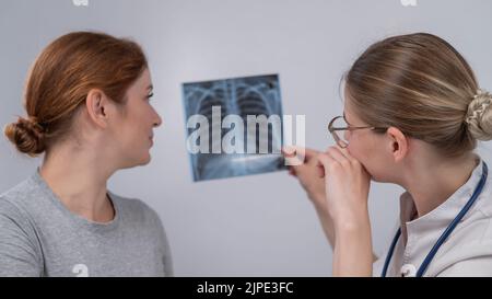 Der Arzt konsultiert eine kaukasische Frau und kommentiert die Röntgenaufnahme der Lunge. Stockfoto