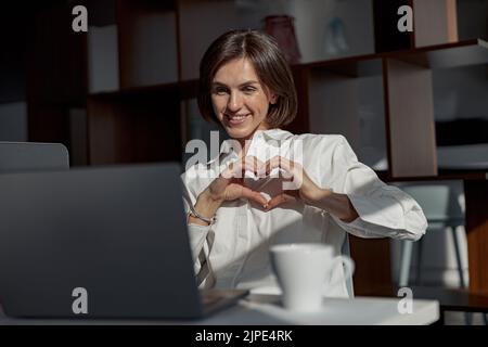 Geschäftsfrau, die im modernen Büro Videoanruf mit Freunden macht und Zeichenliebe zeigt Stockfoto