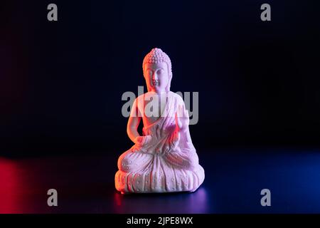 Dekorative weiße Buddha-Statue auf dunklem Hintergrund in rotem und blauem Neonlicht. Dualitätskonzept, Yin Yang. Innerer Kampf der Gegensätze. Wählen Sie Stockfoto