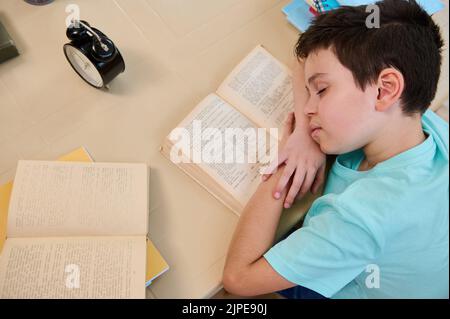 Schuljunge, der auf einem Lehrbuch einschläft und die Müdigkeit bei Hausaufgaben spürt. Schwierigkeiten bei der Wiederherstellung des Schulregimes Stockfoto