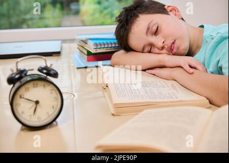 Verschwommener schwarzer Wecker auf dem Hintergrund eines müden schläfrigen Schuljungen, der während der Hausaufgaben auf einem Lehrbuch einschläft. Stockfoto