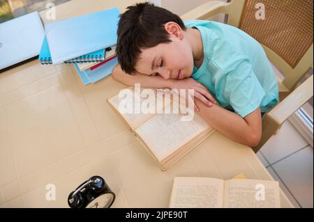 Draufsicht auf einen müden, schläfrigen Schuljungen, der während der Hausaufgaben auf einem Lehrbuch einschläft. Heimschooling. Zurück zur Schule Stockfoto