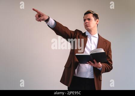 Der Lehrer trägt einen Anzug auf weißem Hintergrund und hält ein Buch, während er irgendwo hinweist. Geschäftsmann zeigt mit einem Buch in der Hand. Anwalt pointin Stockfoto