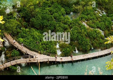 Wandertouristen wandern auf einer hölzernen Fußgängerbrücke, die den See im Naturschutzgebiet umgibt. Majestätischer Blick auf den blauen See inmitten malerischer Landschaft auf den Plitvicer Seen Stockfoto