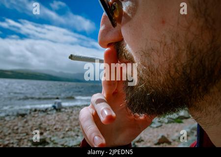 Detail eines Mannes, der ein Marihuana- oder Weed-Joint raucht und sich am Ufer eines Sees entspannt. Stockfoto