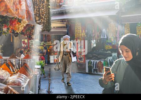 Eine Einheimische, die ihr Telefon überprüft, und ein einheimischer Mann in traditioneller Kleidung, der im Basar durch die Licht- und Rauchstrahlen in Sanliurfa, Türkei, spaziert Stockfoto