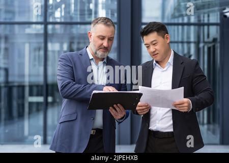 Team aus zwei Geschäftsleuten, die außerhalb des Bürogebäudes arbeiten, diskutieren und überlegen, wie Finanzberichte und -Konten erstellt werden können, einem älteren grauhaarigen Mann und asiatischen Kollegen, Geschäftspartnern Stockfoto