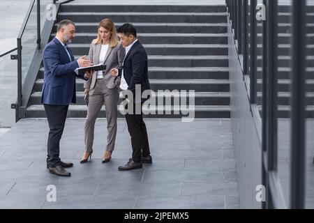 Kollegen, die Mitarbeiter des Unternehmens unterhalten und Geschäftspläne außerhalb des Bürogebäudes besprechen, Männer und Frauen verschiedener Geschäftsteams diskutieren Finanzberichte und Vertragsdokumente auf Treppen Stockfoto