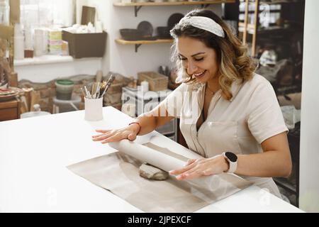 Frau, die Ton rollt, macht keramische Platte im Studio mit Blumenmuster. Handgefertigte kreative Arbeiten. Töpferwerkstatt für Erwachsene. Stockfoto