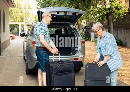 Ältere Ehemann und Ehefrau, die in den Urlaub fahren und Gepäck oder Reisetaschen in den Kofferraum des Autos stecken. Personen, die auf der Pensionstour abreisen, reisen mit Koffern und Trolley im Fahrzeug. Stockfoto