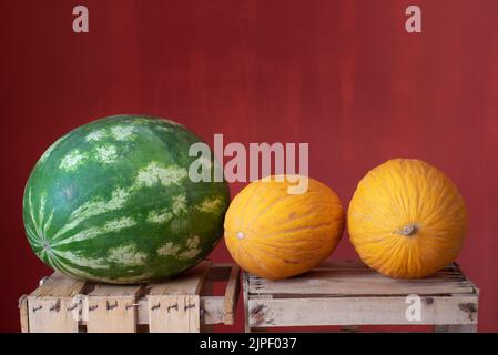 Two yellow ripe honeydew melons and one green watermelon lie side by side on wooden crates for fruit. The background is red. Stockfoto